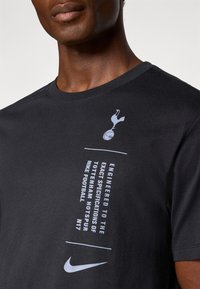 Crna pamučna majica s grafičkim dizajnom, bijelim tekstom i logotipom. Detalji uključuju grb Tottenhama Hotspura i Nikeov znak.