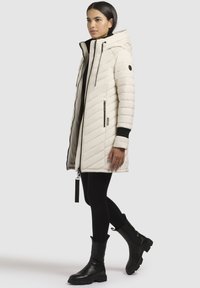 Ung kvinde i en beige, kviltet hættejakke, sorte leggings og sorte ankelstøvler, der står foran en lys baggrund.