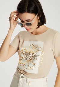Beżowa koszulka z krótkim rękawem z dużym złotym kwiatowym nadrukiem, o gładkiej fakturze. Noszona z dużymi, jasnymi okularami, w swobodnym stylu.