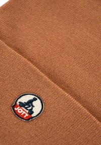 Bruine gebreide stof met een gevouwen rand, met een ronde patch van een persoon die op zijn knieën zit en het woord "JOTT" in rood en wit.