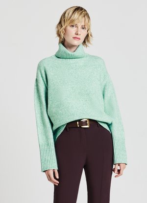 Mintgrüner Rollkragenpullover mit gerippten Bündchen und Saum, in lockerem Schnitt und weicher Textur, kombiniert mit dunkelbraunen maßgeschneiderten Hosen.