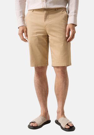 Mężczyzna ubrany w beżowe szorty do kolan, jasnobarwną koszulę z długim rękawem oraz beżowe klapki z czarnymi podeszwami, stojący na białym tle.