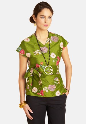 Frau trägt eine grüne, geblümte Wickelbluse mit großer Schleife, schwarze Hose, eine lange schwarze Perlenkette mit rundem Anhänger und goldene Armreifen.