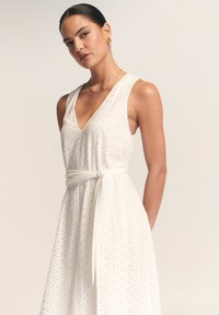 Balta suknelė be rankovių su V formos iškirpte, pasižyminti skylučių raštu, surištu liemens diržu ir laisvai krentančia suknele. Lygus tekstūra ir elegantiškas dizainas.