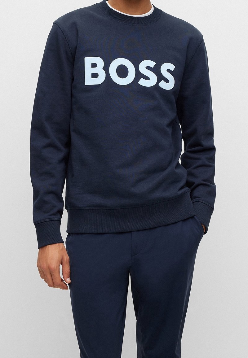 Homme portant un sweat-shirt bleu marine avec de grandes lettres blanches formant "BOSS" sur la poitrine et un pantalon bleu marine assorti.