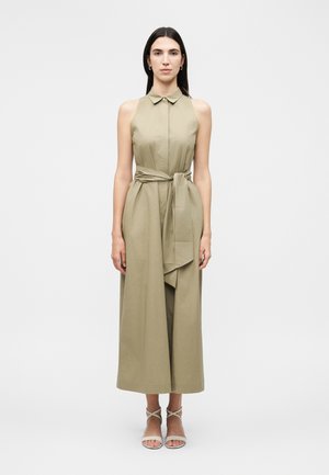 Nő áll, ujjatlan olívazöld galléros midi ruha derékszalaggal és bézs pántos magassarkú szandál viselése egyszerű háttér előtt.