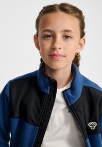 Blauwe en zwarte fleecejas met een opstaande kraag, ritssluiting en logopatch. Het ontwerp bevat kleurvlakken en een zachte textuur.