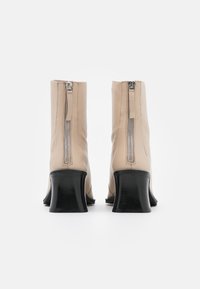Stivaletti alla caviglia in pelle beige con zip argento, tacco nero curvo basso e una texture liscia. Visuale posteriore che mette in evidenza il dettaglio della zip.