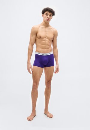 Męski model stojący boso, ubrany w fioletowe bokserki Polo Ralph Lauren na jednolitym białym tle.