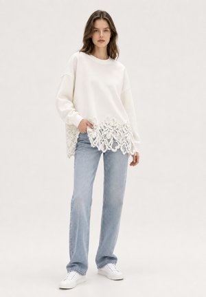 Kvinde iført hvid langærmet top med blondekant, lyseblå jeans og hvide sneakers, stående med den ene hånd i lommen mod ensfarvet baggrund.