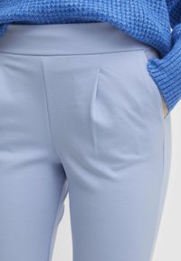 Helles, blaugraues, maßgeschneidertes Paar Hosen mit glatter Textur, ausgestattet mit vorderen Falten und seitlichen Taschen. Kombiniert mit einem grob gestrickten blauen Pullover.