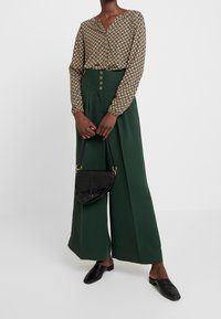 Langärmelige, gemusterte Bluse in Beige und Braun, kombiniert mit hochtaillierten, grünen, weiten Hosen mit goldenen Knöpfen; hält eine schwarze Handtasche.