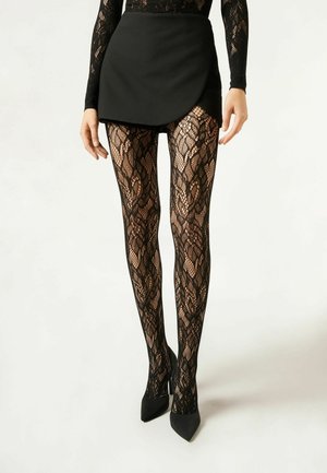 Collant in pizzo nero con un motivo floreale, abbinati a uno skort nero e tacchi alti a punta, mostrano un design strutturato.