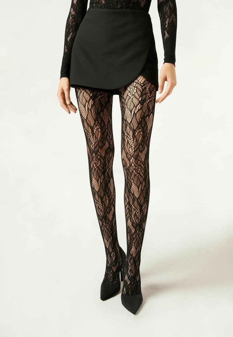 Collants en dentelle noire avec un motif floral, associés à un short skort noir et des talons aiguilles, mettent en valeur un design texturé.