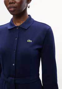 Polo di colore blu navy realizzato in tessuto testurizzato, con colletto, chiusura a bottone e un piccolo logo di coccodrillo verde sul petto. Fissato in vita con una cintura.