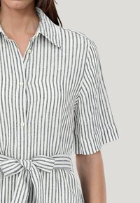 Camicia a maniche corte con bottoni a righe verticali bianche e navy, caratterizzata da una vita legata e da un colletto classico. Tessuto leggero con superficie liscia.