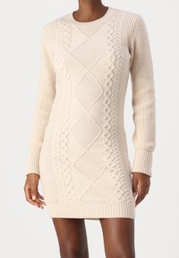 Robe pull en tricot beige présentant des motifs en relief en câble, des poignets côtelés et un col rond. Longueur mi-cuisse et silhouette ajustée.