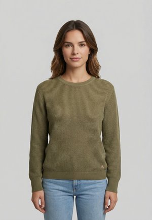 Mujer con cabello castaño hasta los hombros, vestida con un suéter de punto verde oliva y jeans azul claro, de pie frente a un fondo gris liso.