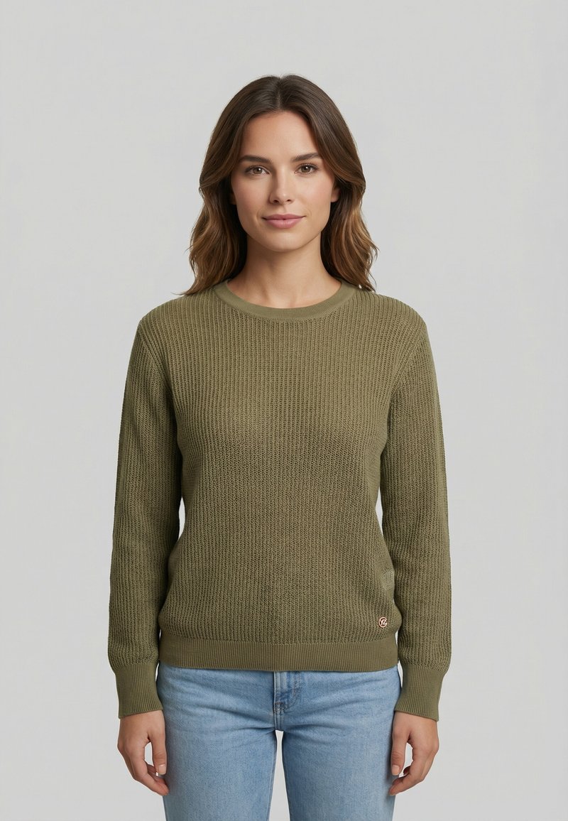 Donna con capelli castano chiaro fino alle spalle, indossa un maglione a maglia verde oliva e jeans azzurri, in piedi davanti a uno sfondo grigio uniforme.