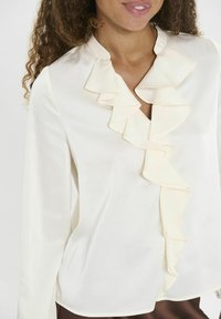 Crème blouse met lange mouwen en een ruches halslijn. De gladde stof en subtiele glans benadrukken het gestructureerde ontwerp en de elegante details.