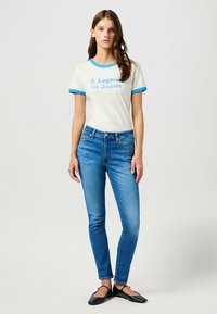 Vit T-shirt med blå ringkrage och trycket "A Legend in Jeans"; slim fit blå denimjeans; svarta platta skor; slät tyg.