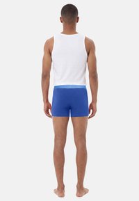Man standing barefoot facing away wearing a white tank top and blue fitted boxer briefs against a plain light background.