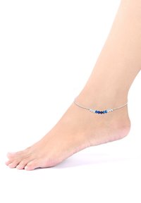 Silbernes Knöchelarmband mit blauen und klaren Perlen in verschiedenen Formen, das auf einem nackten Fuß vor einem weißen Hintergrund liegt.