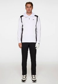 Vit halvlång dragkedjepullover med svarta sidodetaljer, kombinerad med svarta byxor och en vit handske. Innehåller en logotyp på bröstet och sportiga skor.