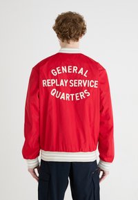 Red bomber jacket with white text on back: “GENERAL REPLAY SERVICE QUARTERS.” Features ribbed white cuffs and waistband. Smooth fabric texture.