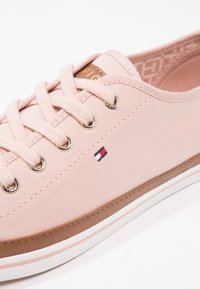 Baskets en toile rose clair avec semelle blanche et marron, lacets roses, œillets métalliques et petit logo rectangulaire rouge, blanc et bleu sur le côté.