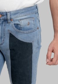 Jeans in denim azzurro chiaro con un pannello in tessuto scuro sulla coscia, dettagli in rame e cuciture a contrasto.