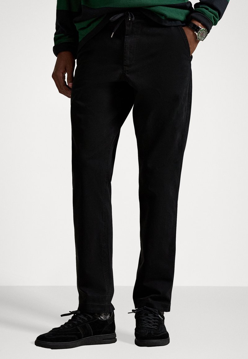 Black cotton trousers with a relaxed fit, featuring a drawstring waist, side pockets, and a smooth, matte texture. Paired with black sneakers.