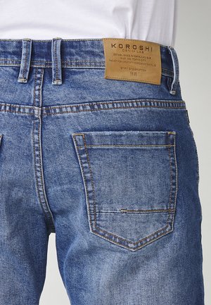 Blue denim jeans with a leather brand patch on the back waistband, five-pocket design, and contrast stitching. Lightly worn appearance.