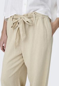 Béžové lněné kalhoty s uvolněným střihem, elastickým pasem a uvázaným páskem. Obsahují boční kapsy a hladký, texturovaný povrch.
