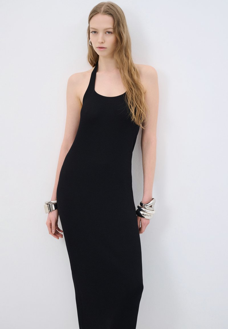 Abito nero a costine con una silhouette aderente. Il modello indossa braccialetti in argento sovrapposti e si trova di fronte a uno sfondo semplice.