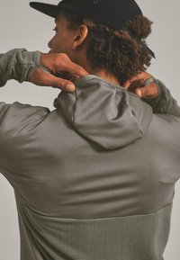 Person mit lockigem Haar in einer schwarzen Mütze zieht die Kapuze eines grau strukturierten Hoodies hoch, von hinten gegen einen neutralen Hintergrund gezeigt.