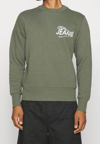 Sudadera verde oliva hecha de una mezcla de algodón, con cuello redondo, puños y dobladillo acanalados, y texto gráfico blanco en el pecho.