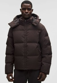 Ruskea puhallettava takki korkealla kauluksella, vetoketjukiinnityksellä ja irrotettavalla hupulla. Sisältää tikattuja osia ja kaksi sivutaskua.