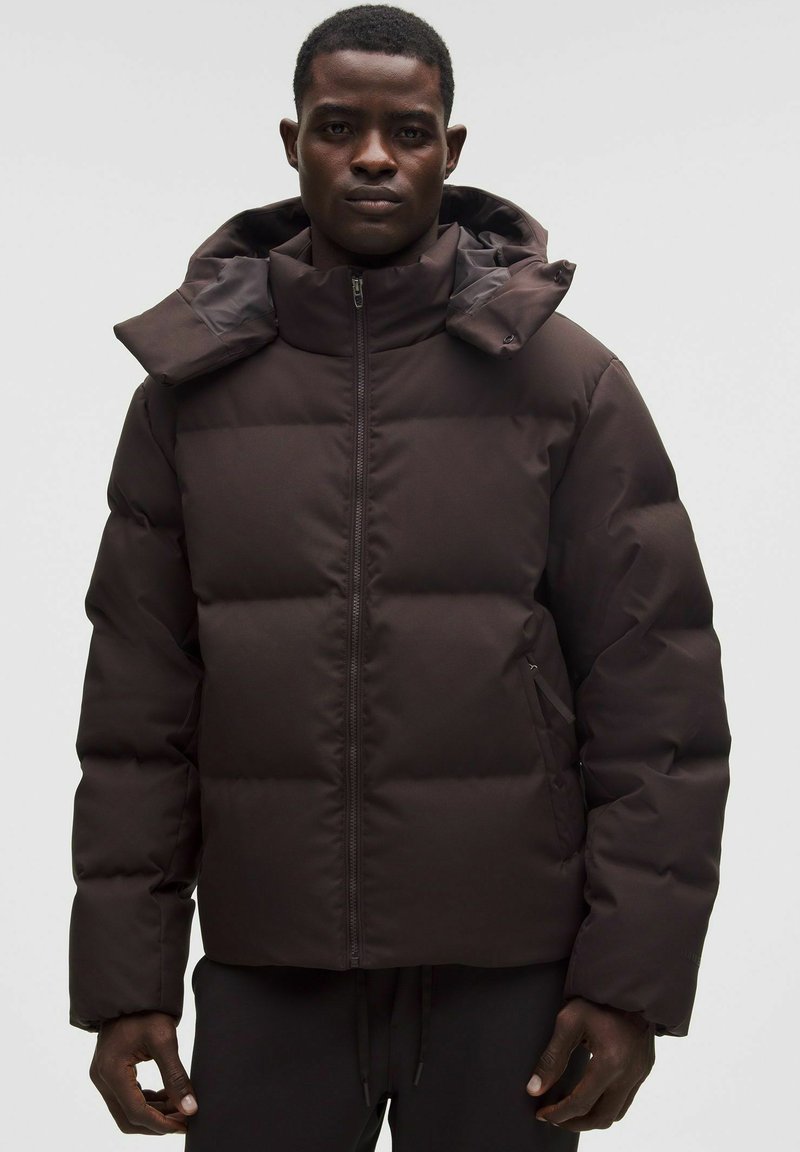 Ruskea puhallettava takki korkealla kauluksella, vetoketjukiinnityksellä ja irrotettavalla hupulla. Sisältää tikattuja osia ja kaksi sivutaskua.