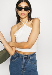Top cropped branco com alças finas, combinado com jeans azuis de cintura alta. Os acessórios incluem óculos de sol pretos em formato de gato e uma bolsa verde.