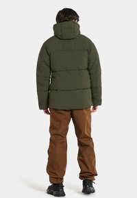 Doudoune verte avec capuche, dotée de sections matelassées. Associée à un pantalon marron et des chaussures noires. Design simple et fonctionnel.