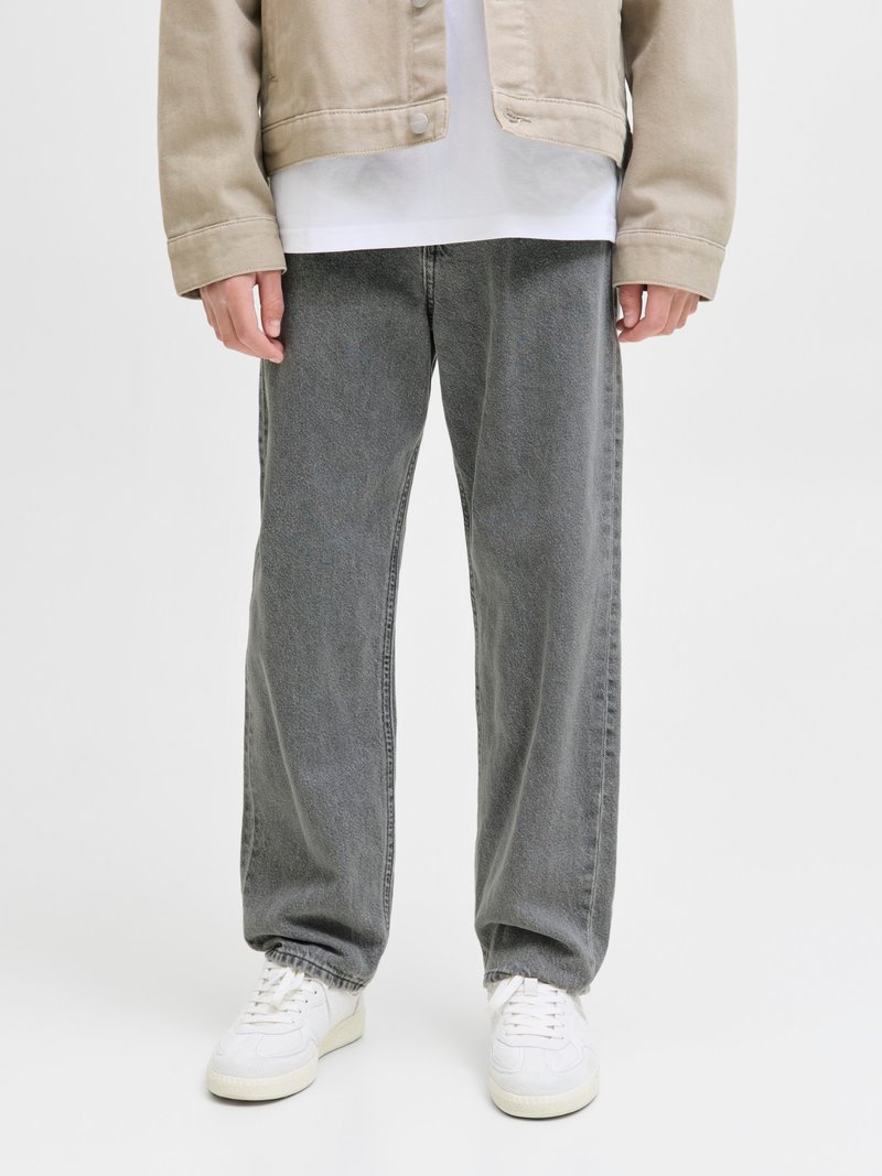 Person som har på seg løse grå jeans, hvite joggesko, beige jakke og hvit skjorte, står mot en ensfarget lys bakgrunn.