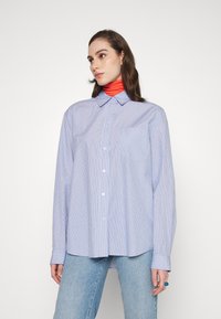 Camisa de botão listrada em azul e branco, com mangas longas e colarinho; apresenta um bolso e um ajuste descontraído. Usada sobre um suéter de gola alta vermelho.