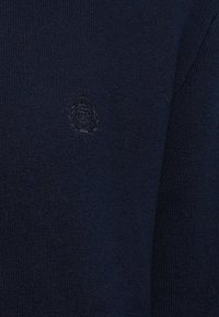 Pulover tricotat albastru marin, cu un guler rotund și un emblema brodat subtil pe piept. Textură moale, cu un finisaj ribetat.