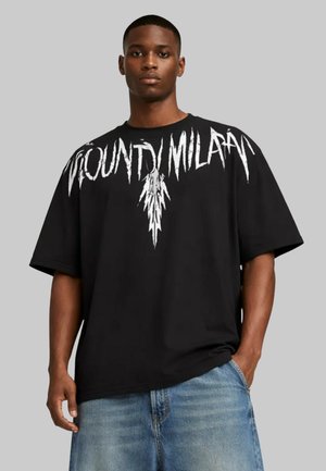 Giovane uomo che indossa una t-shirt nera oversize con testo bianco dentellato "WOUND MILANO" sul petto e jeans azzurri chiari.