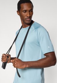 Αθλητική μπλούζα ανοιχτού γαλάζιου με στρογγυλή λαιμόκοψη και κοντά μανίκια. Φέρει μικρό λογότυπο στο στήθος και υφασμάτινη υφή. Κρατάει σχοινάκι άλματος.