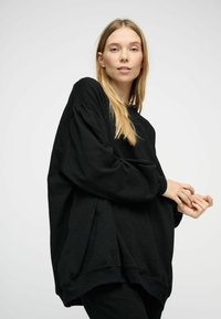 Noor naine, kellel on pikad blondid juuksed, kannab laia musta dressipluusi ja poseerib käed koos lihtsa heleda halli tausta ees.