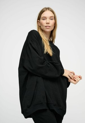 Giovane donna con lunghi capelli biondi che indossa un ampio felpa nera, posa con le mani unite su uno sfondo grigio chiaro uniforme.