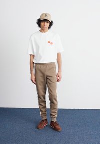 Camiseta blanca de manga corta con un gráfico en rojo y naranja, combinada con pantalones de chándal marrón claro y zapatos marrones. Una gorra color beige completa el conjunto.