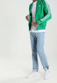 Giacca verde con zip e strisce bianche, indossata sopra una maglietta bianca. Jeans azzurri chiari e sneakers bianche completano l'outfit.